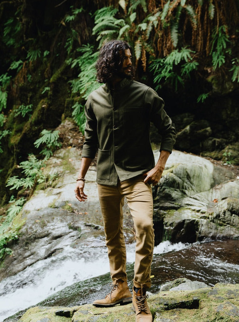 Full-length view of the Tambo Wool Jacket in Olive Green with khaki trousers and leather boots near a waterfall