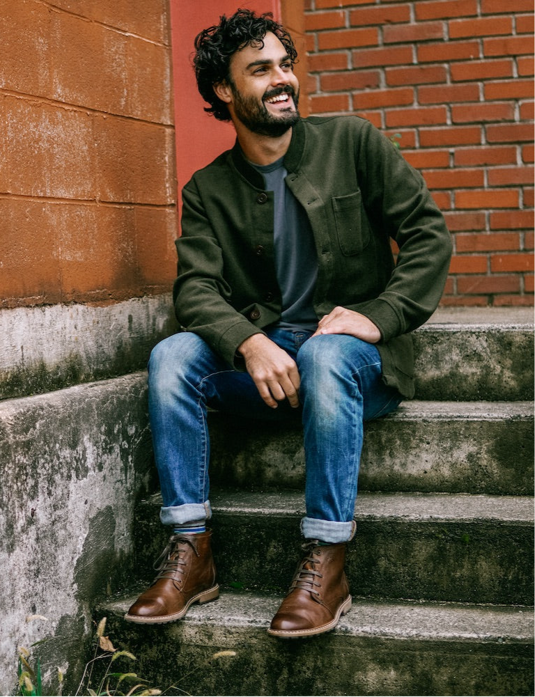 Smiling man sitting on stone steps in the Tambo Wool Jacket in Olive Green, with jeans and brown boots