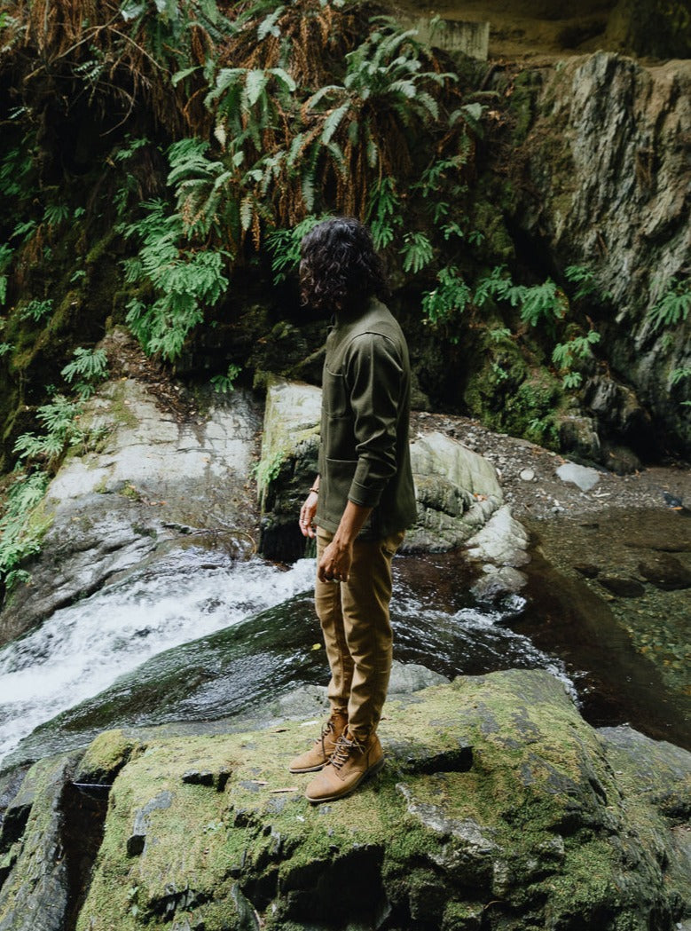 Side view of a man on mossy rocks by a waterfall in the Tambo Wool Jacket in Olive Green